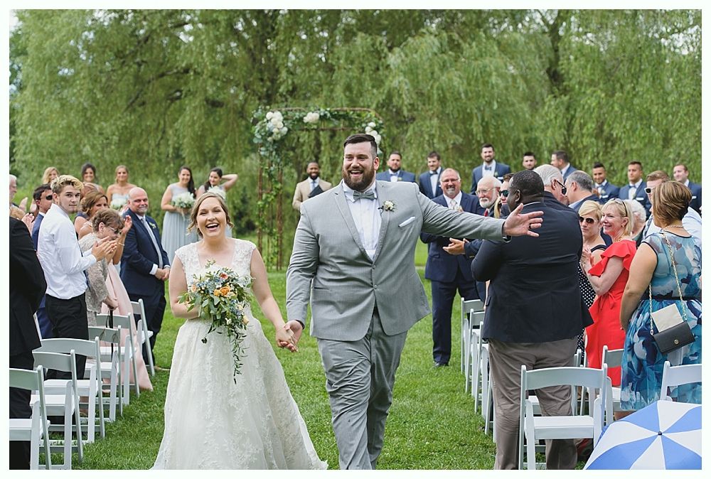 Newlyweds walk down aisle, smiling. Wedding guests cheer. Outdoor ceremony.