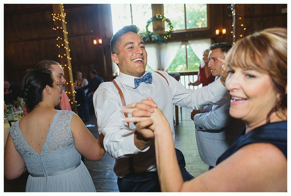Man dancing with a woman at a wedding reception; string lights hang, wooden beams in background.