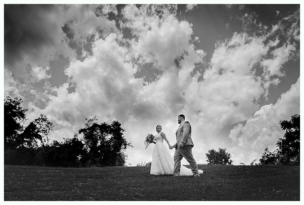 Bride and groom holding hands, walking on a grassy field, cloudy sky in background. Black and white.
