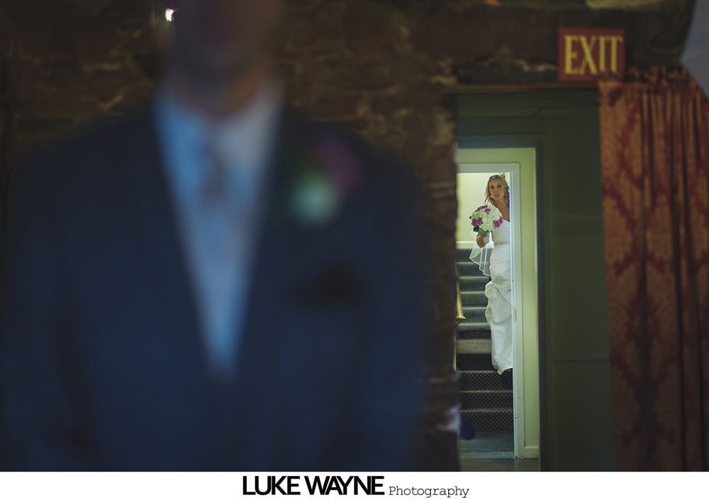 Groom blurred in foreground. Bride in white dress and bouquet appears in doorway.