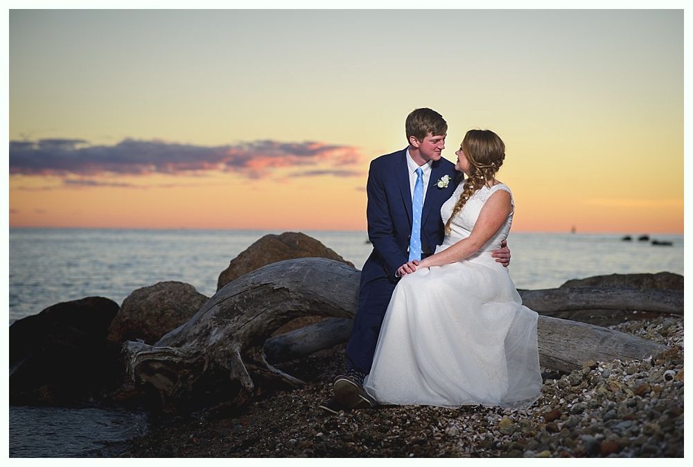 Bride and groom embrace on rocks by the ocean at sunset. The groom wears a blue suit, the bride a white dress.