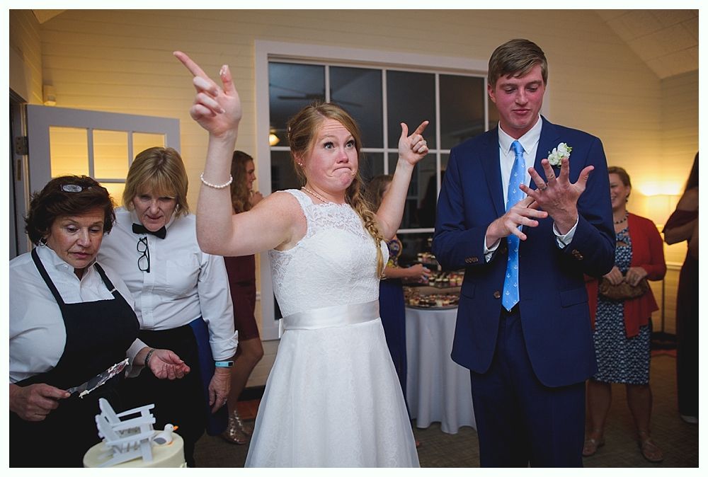 Bride with arms up, making a funny face, groom looking at his hand, cake cutting in background.