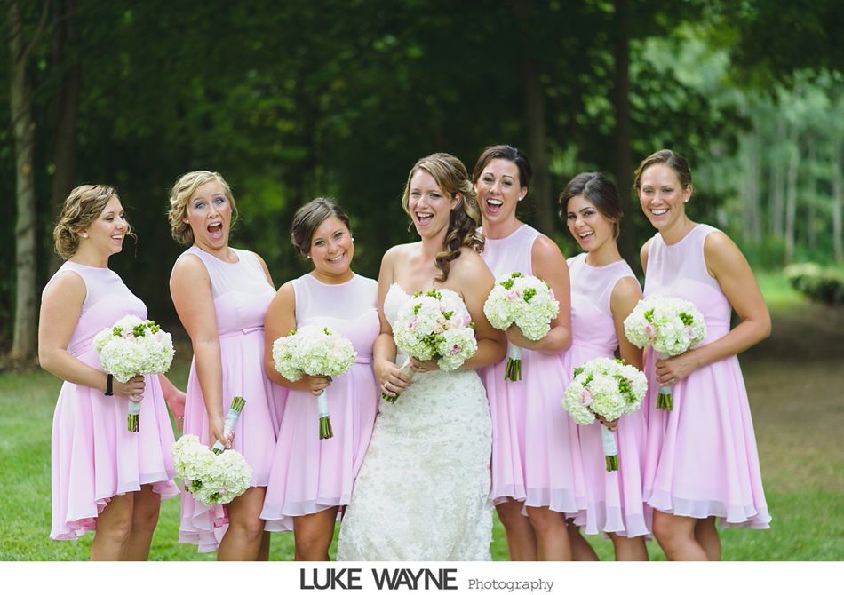 Bride and bridesmaids in pink dresses, laughing and holding bouquets in a green outdoor setting.