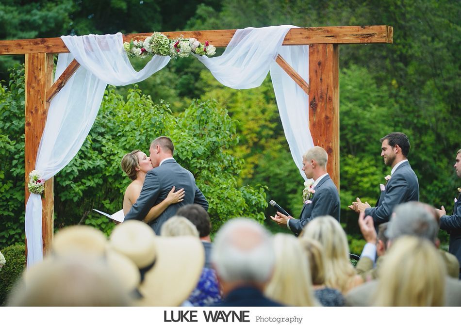 Couple kissing at outdoor wedding ceremony under a draped wooden arch; guests seated.