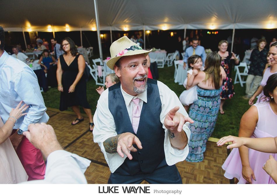 Man dancing enthusiastically at an outdoor wedding reception, wearing a hat, surrounded by other guests.