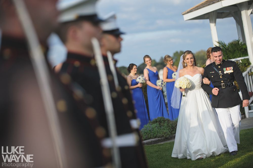 Bride and groom walk down sidewalk, groom smiles, bride holds purple bouquet, city background.