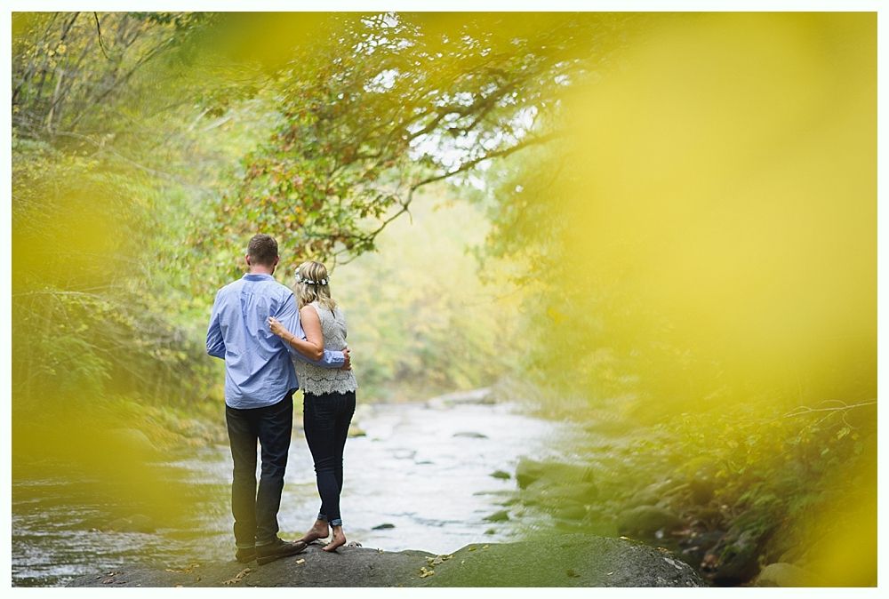 Couple standing by a stream, arms around each other, framed by blurred yellow foliage.