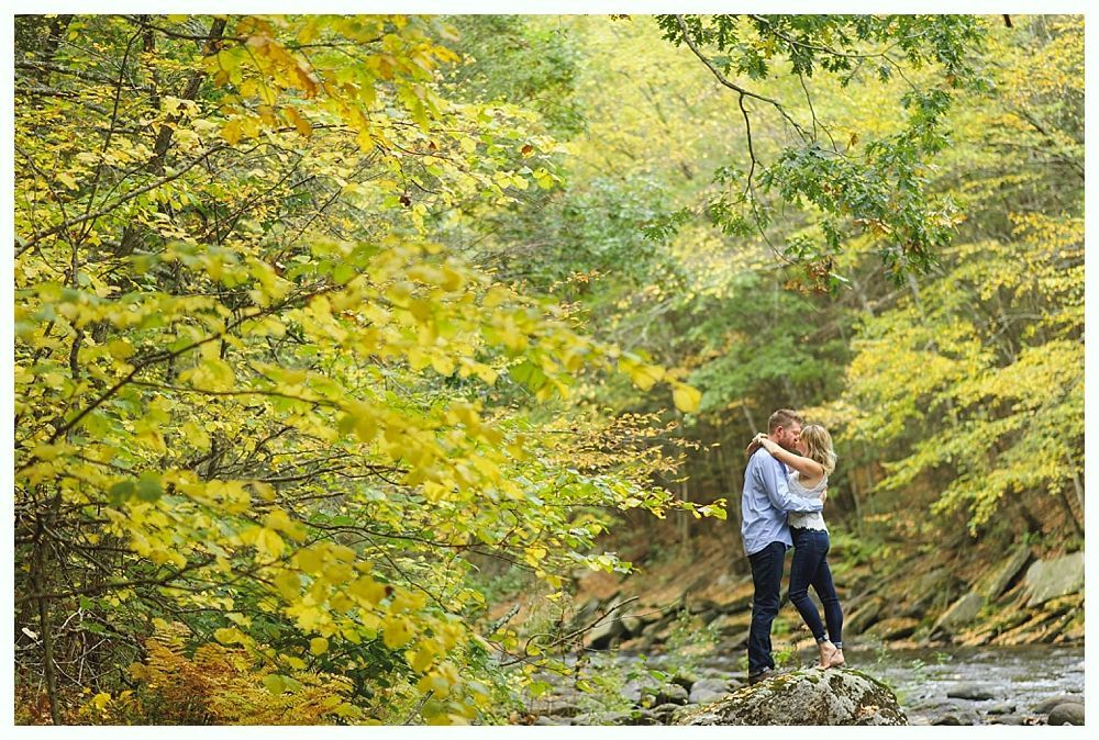 Couple kissing near stream in autumn foliage. Yellow and green leaves surround them.