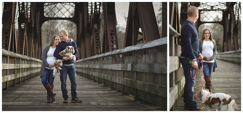 Pregnant couple and small dog pose on a bridge. The man holds the dog while the woman cradles her belly.