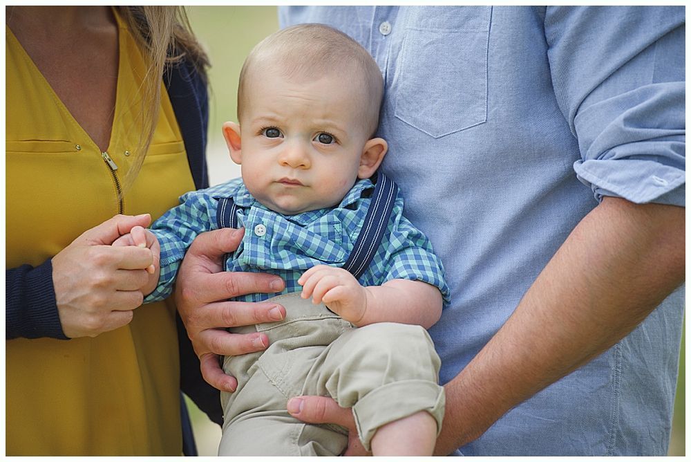 Baby held by two adults, baby wearing suspenders and blue checkered shirt.