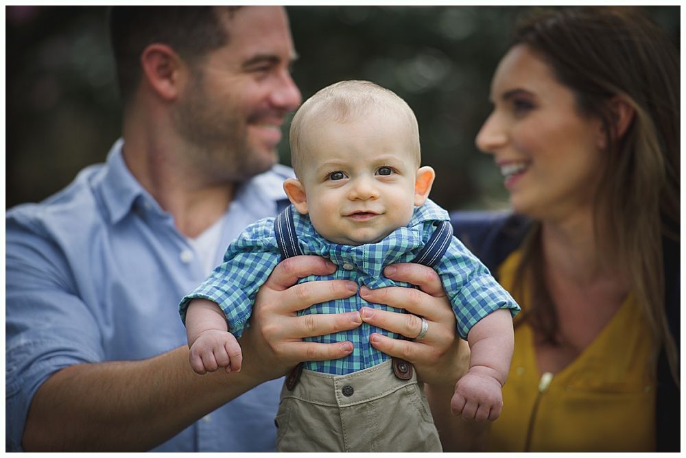 A baby being held by a smiling man and woman outdoors. Baby smiles, wearing suspenders.