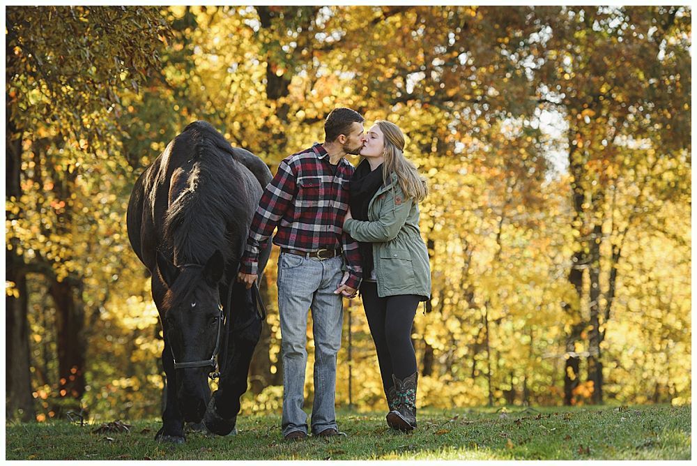 Couple kissing near a black horse in an autumn field. Man wears plaid, woman a green jacket.