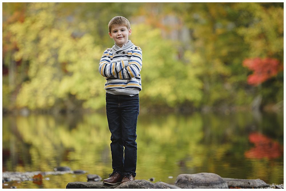 Boy with arms crossed standing on rocks; fall foliage backdrop.