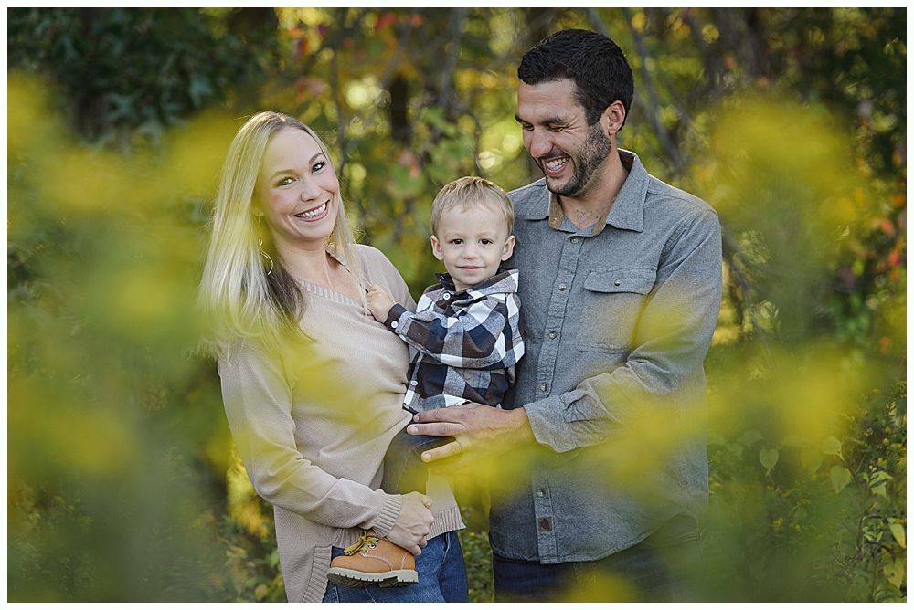Family of three smiling outdoors, foliage in foreground.