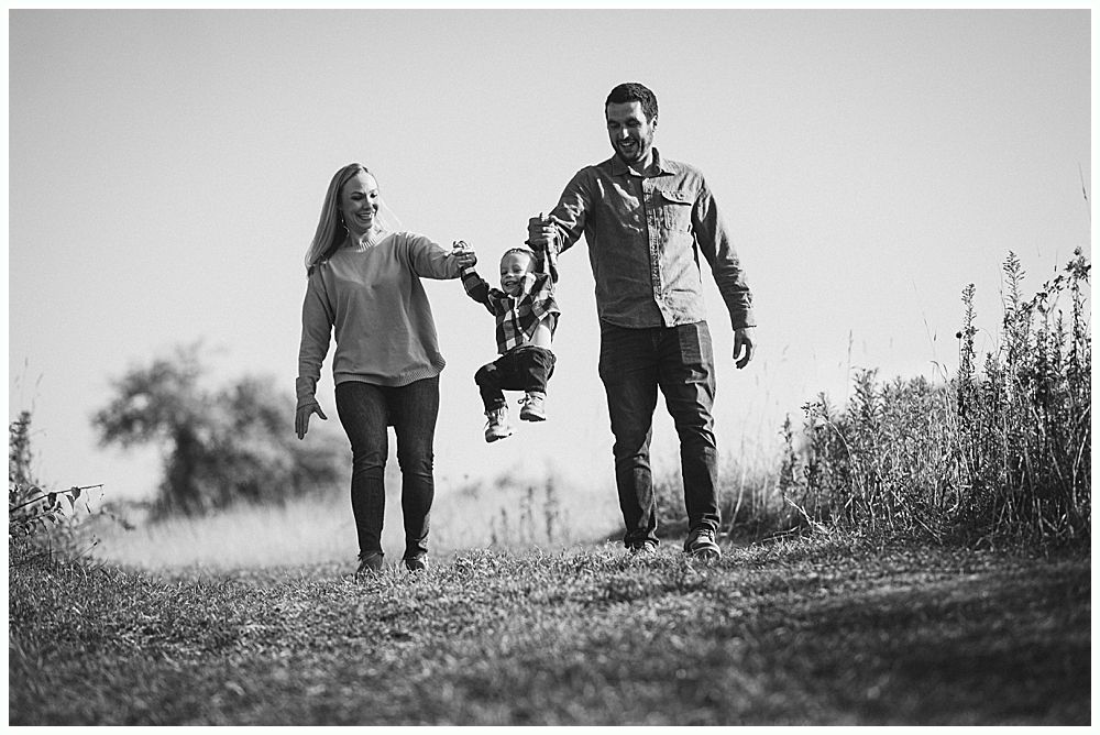Family walking, parents holding a child suspended in the air. Grassy path, bright sky. Black and white.