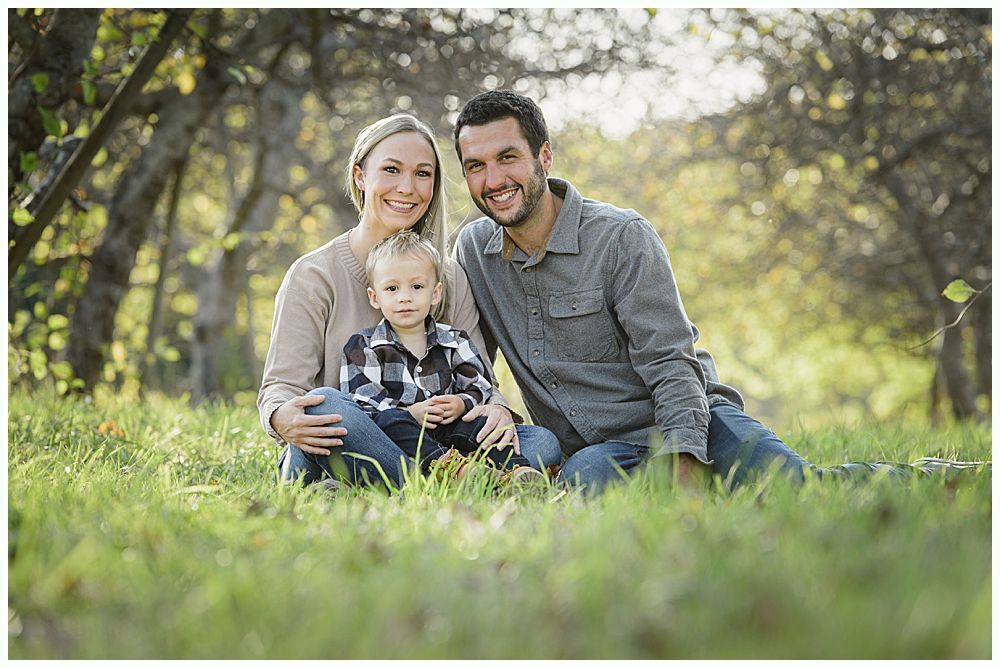 Family of three smiling, sitting in a grassy field under a tree canopy.