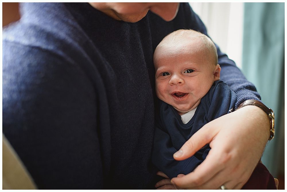 Smiling baby held by a person wearing a blue sweater; indoors, natural light.