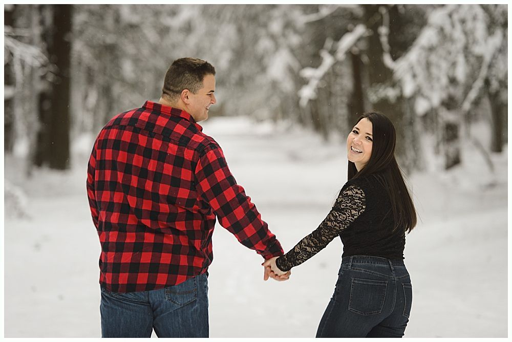 Couple holding hands walking in a snowy forest. Man in red plaid shirt smiles, woman laughs, looking over shoulder.