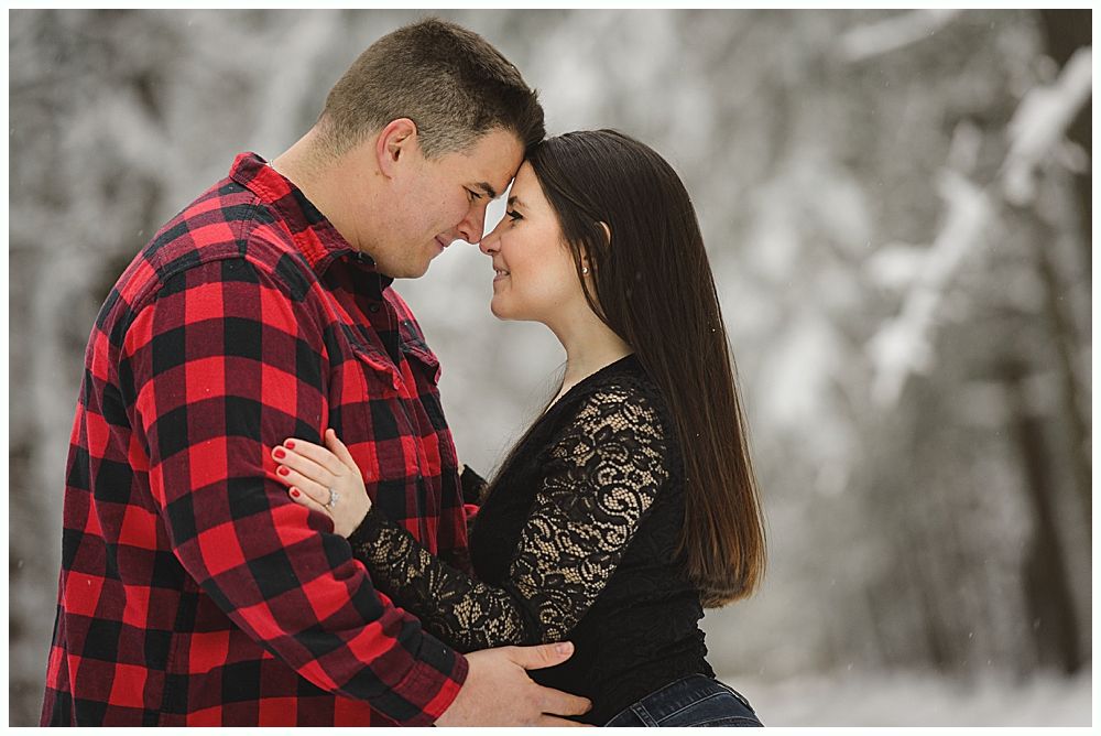 Couple gazing at each other forehead to forehead, standing in snow-covered woods; man in red plaid, woman in black lace.