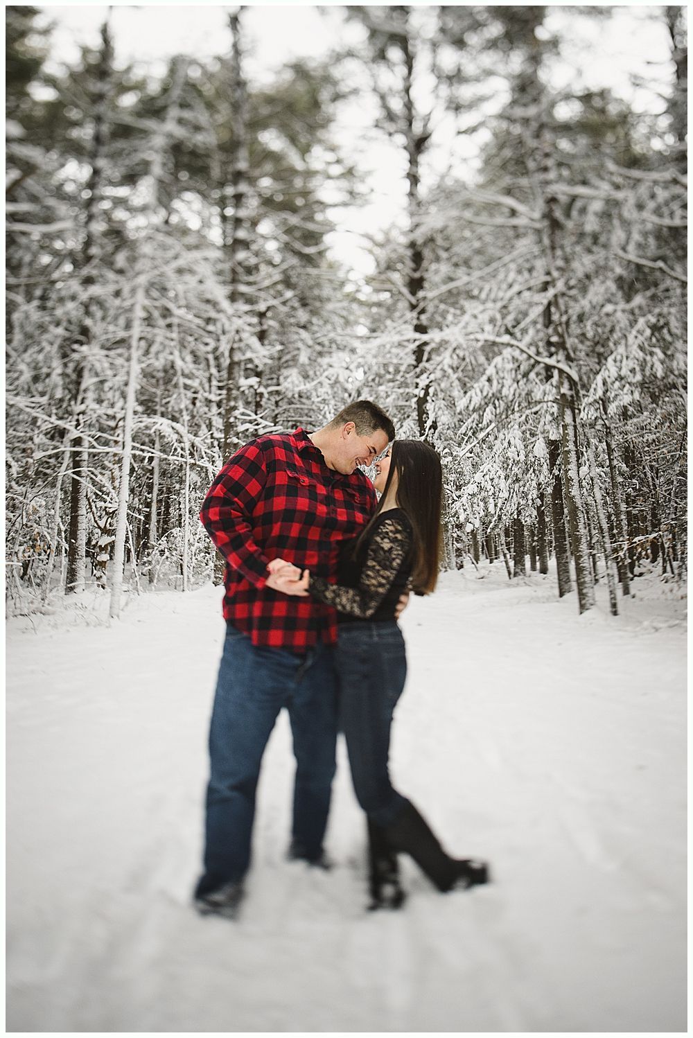Couple kissing in a snowy forest; man in plaid shirt, woman in black lace.
