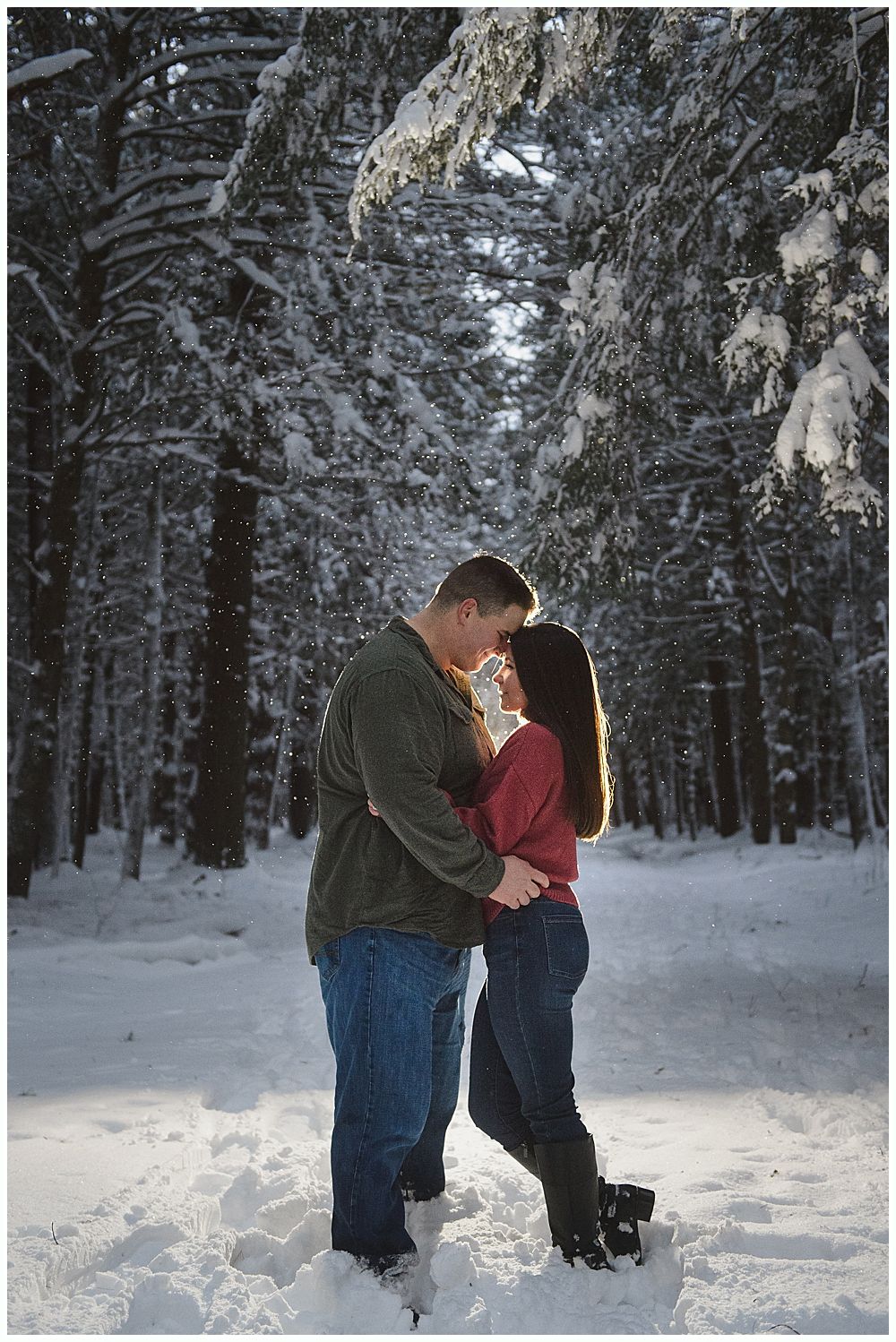Couple embracing in snowy forest; sunlight shines through trees.