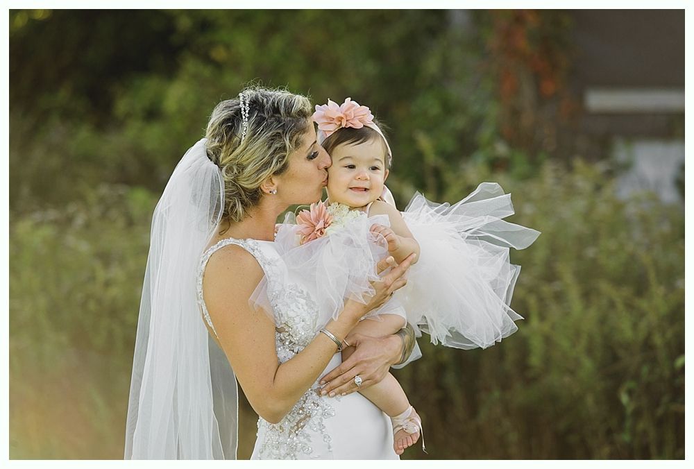 Bride in a wedding dress kisses a baby wearing a matching dress and flower headband. Outdoors with greenery.