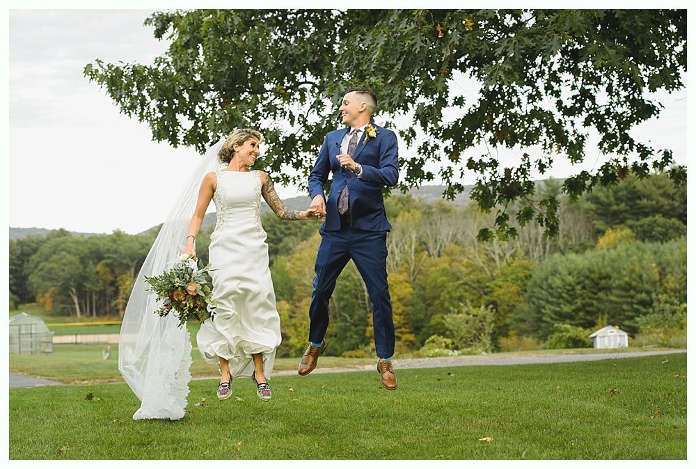 Bride and groom jump in the air, holding hands. Outdoors in a field under a tree, fall foliage visible.