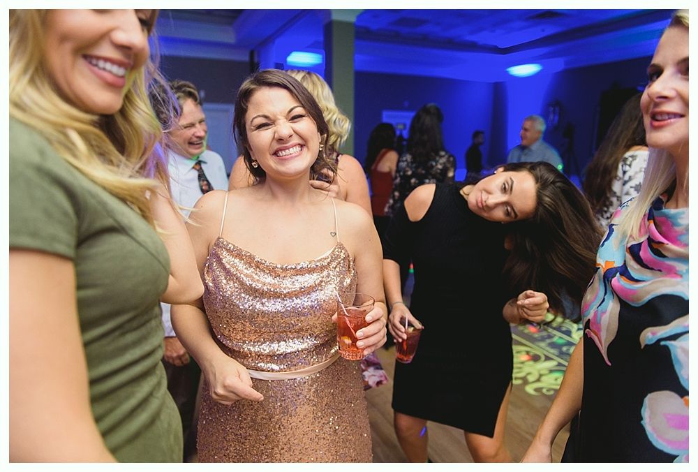People dancing at a party, smiling, holding drinks. Room with blue lighting, people in the background.