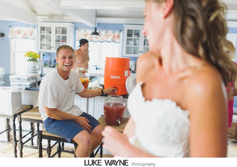 Man smiles at bride in kitchen, filling a pitcher from a dispenser. Others in background.