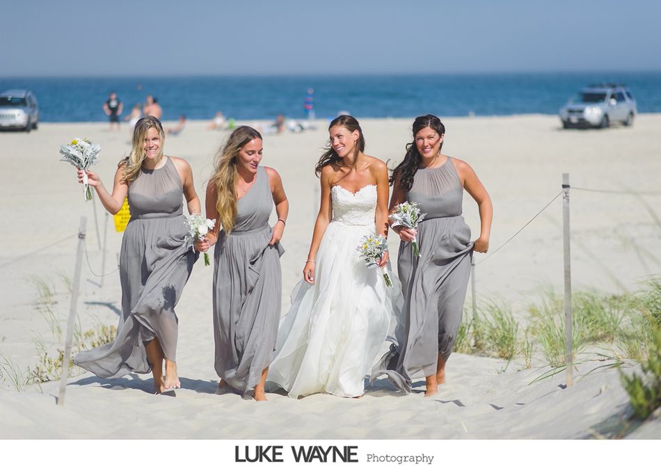 Bride and bridesmaids walk on a beach, wearing a white wedding dress and gray gowns. Sunny day.