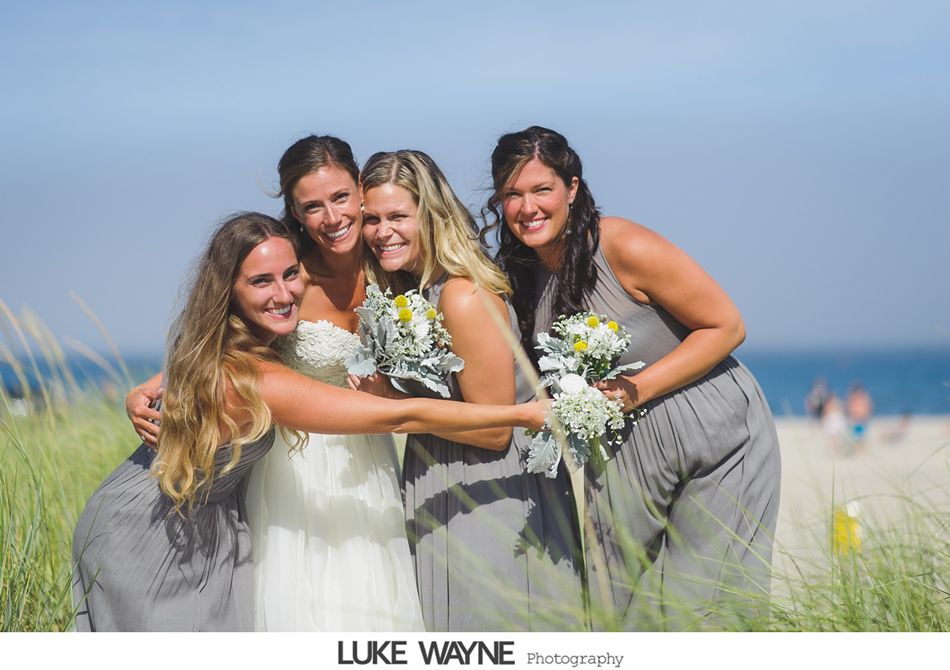 Bride and bridesmaids embrace on a sunny beach, holding bouquets. Gray dresses, blue sky, and ocean in the background.