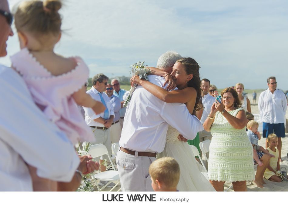 Bride hugs person at beach wedding ceremony. Guests watch and photograph.
