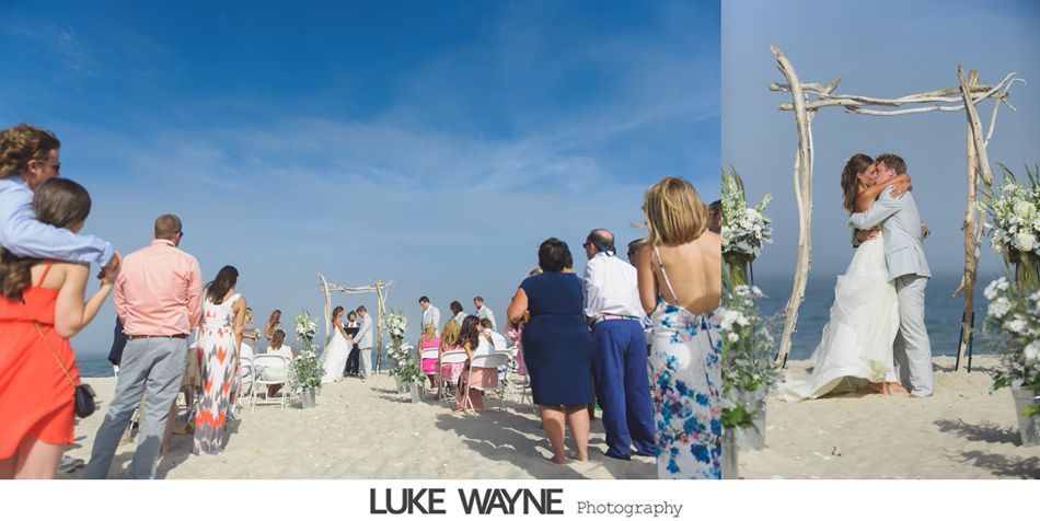 Beach wedding with ceremony under driftwood arch; couple kissing, guests watching.