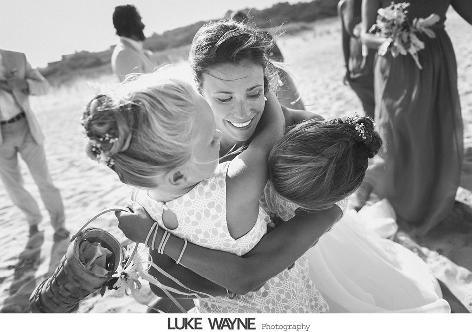Bride hugs two flower girls on a sandy beach; all are smiling.