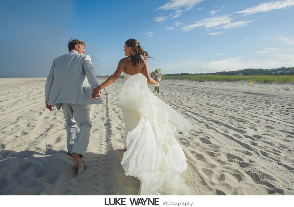 Newlyweds walk hand-in-hand along a sandy beach. She wears a white gown, and he wears a light-colored suit.