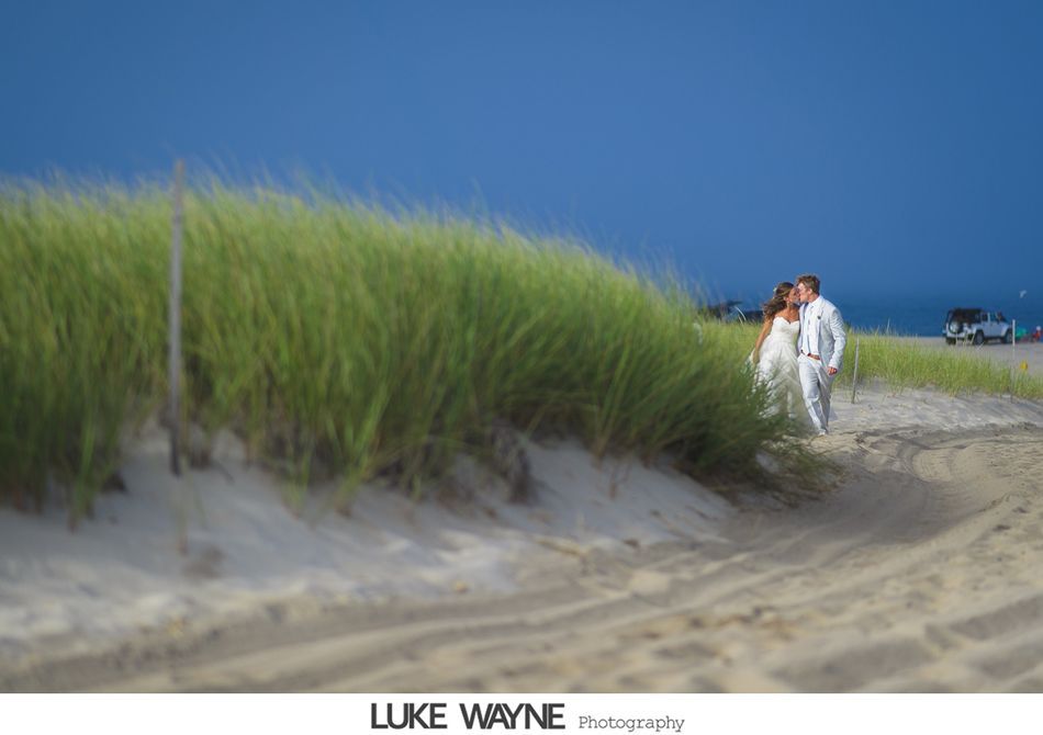 Bride and groom kiss on a sandy path, with tall green grass, blue sky, and ocean.