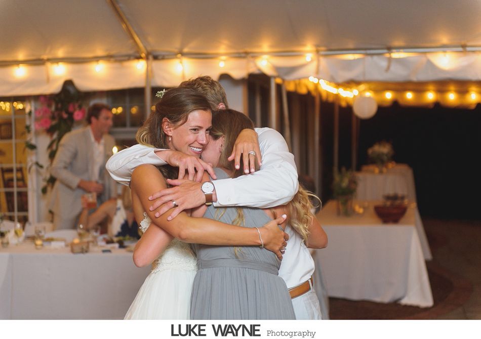 Bride hugs two women under a tent, smiling. Wedding reception, lights overhead.