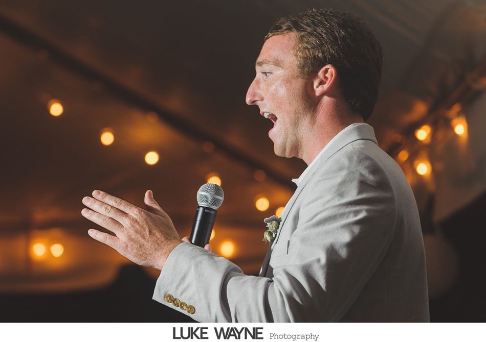 Man in a light-colored suit speaks into a microphone at a wedding reception, gesturing with his hands; warm lights in the background.