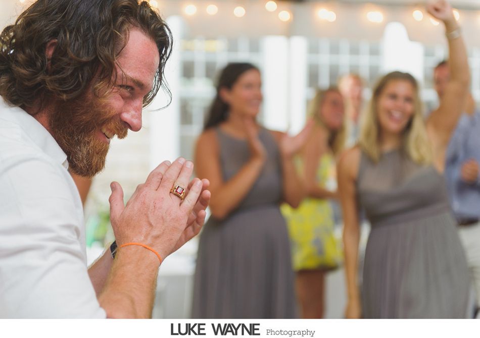 Man clapping, laughing at a gathering. Women in gray dresses dancing in the background. Festive lighting.