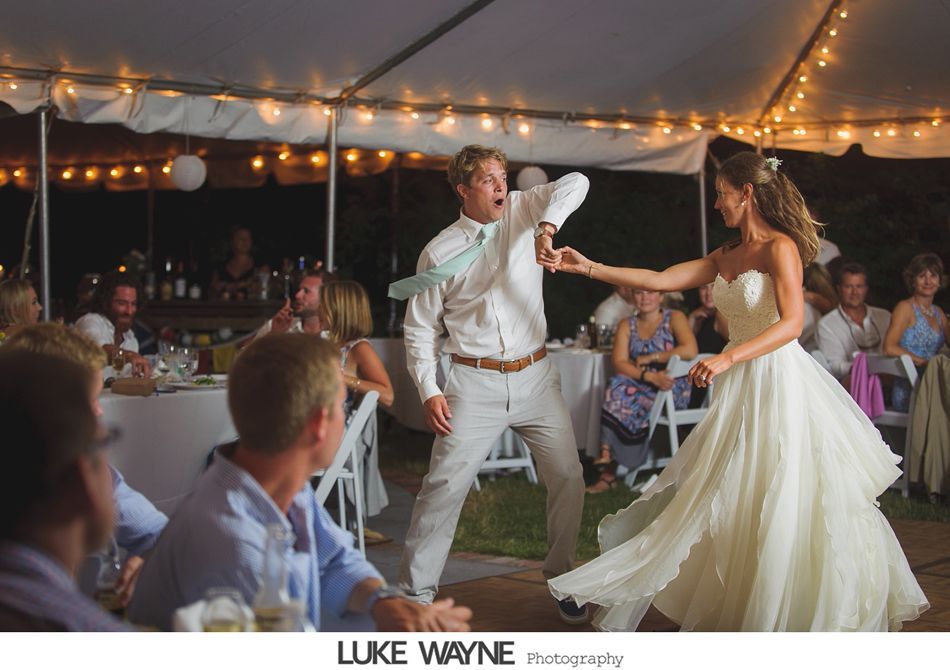 Groom dances with bride at a wedding reception under string lights. Both are smiling, he is mid-motion with his arms raised.