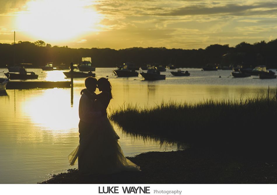 Couple silhouetted by sunset over water with boats.