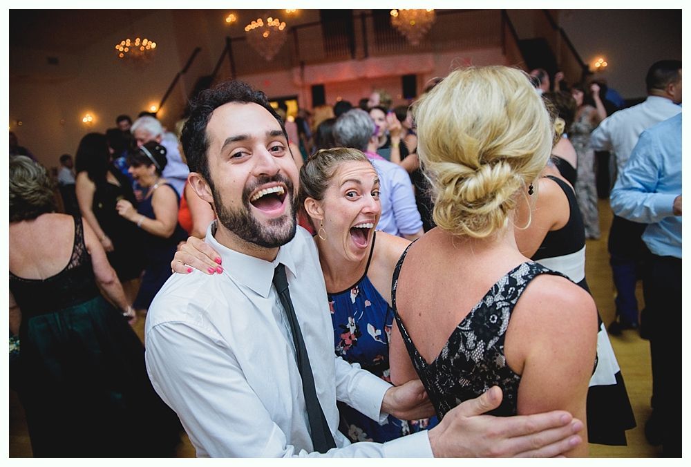 Bride with arms up, making a funny face, groom looking at his hand, cake cutting in background.