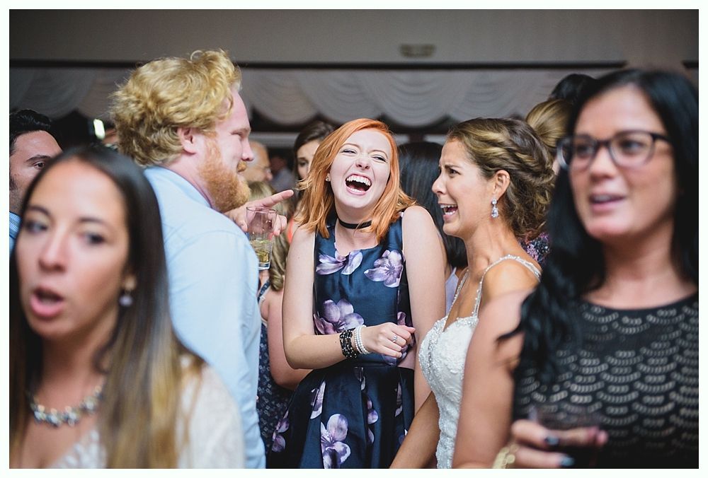 Bride with arms up, making a funny face, groom looking at his hand, cake cutting in background.