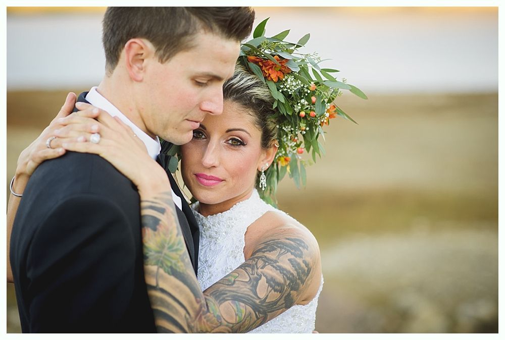 Bride and groom embrace. Bride wears white lace dress, floral crown, and has arm tattoos. Groom in black suit.