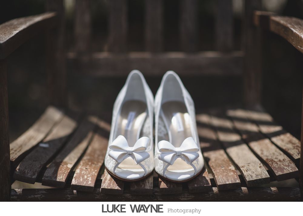 White bridal heels with bow detail, resting on a weathered wooden chair.