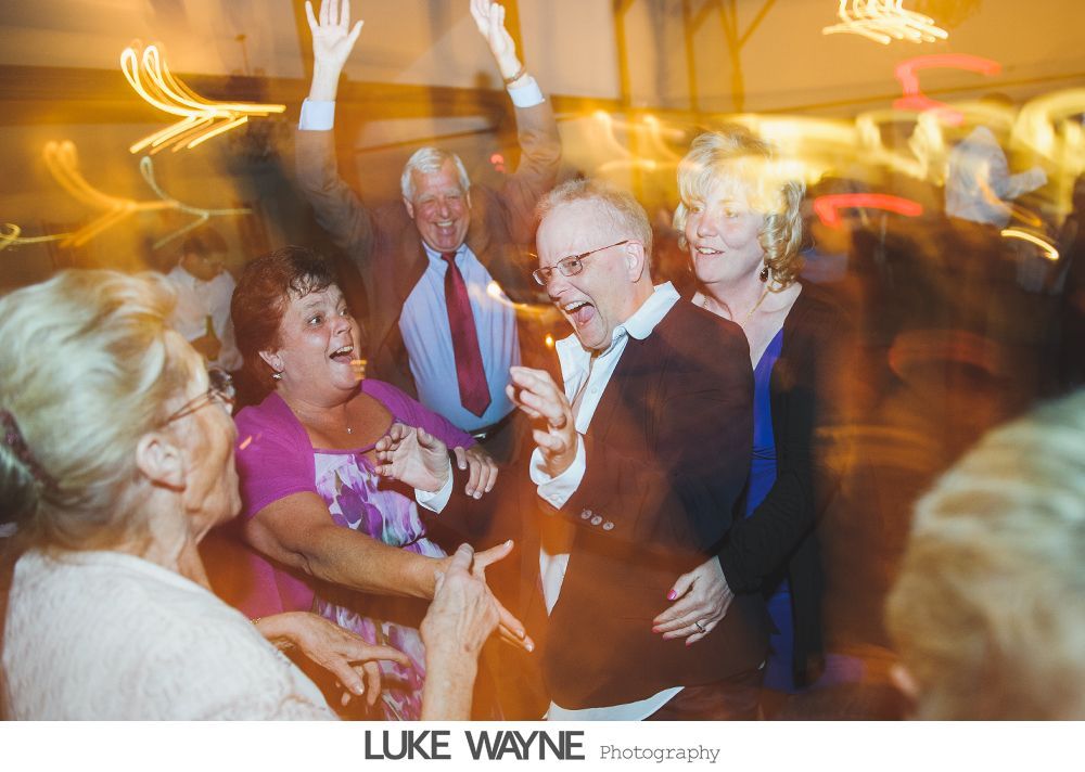 People dancing joyfully at an event with blurred lights; a man in a blazer laughs amidst the movement.