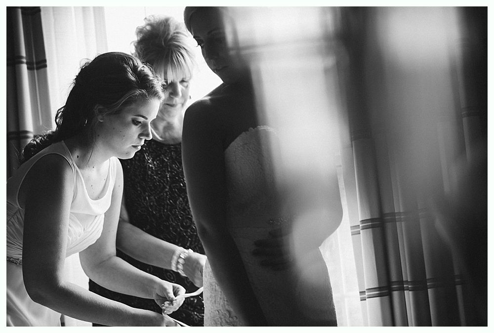 Woman in wedding dress being helped by two others, near window.
