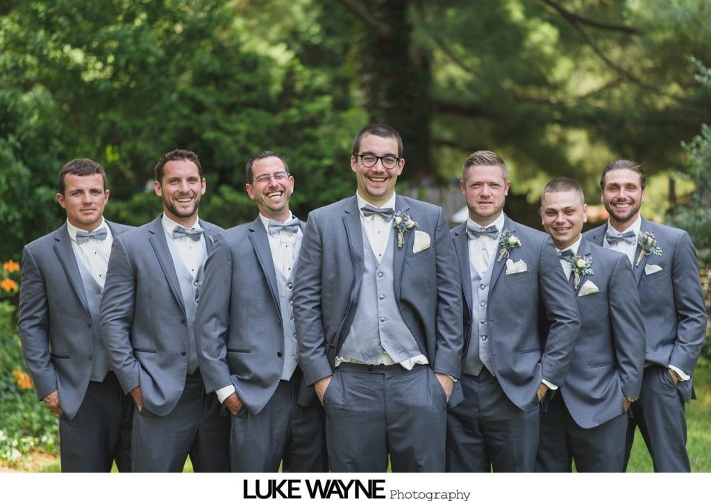 Seven men in gray suits pose outdoors; some smiling, some with hands in pockets.