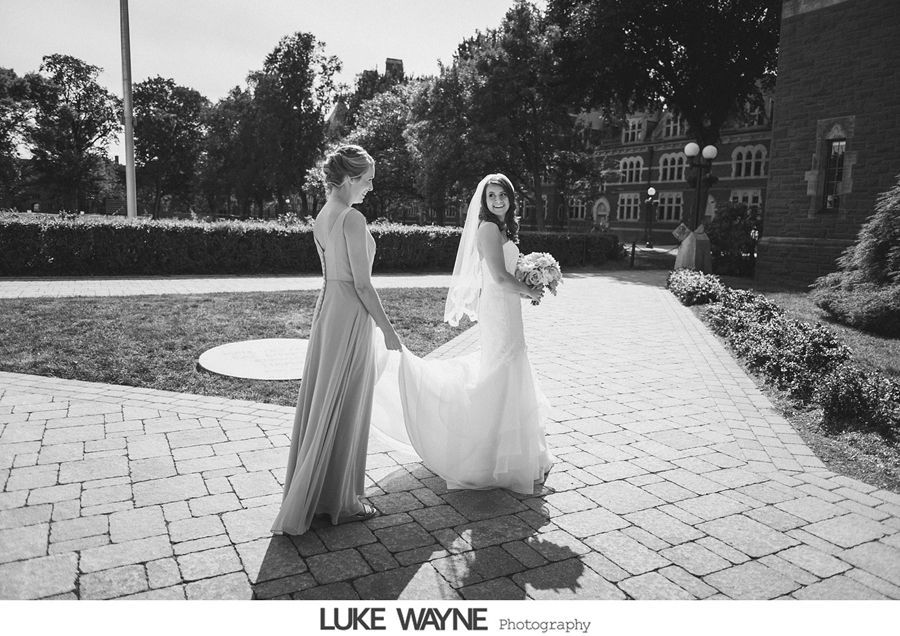 Bride and bridesmaid walking on brick path, holding hands, sunny day.