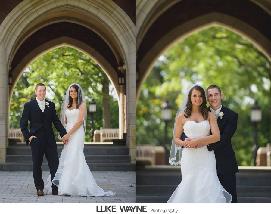 Bride and groom pose under an arched walkway; the groom wears a suit and the bride, a strapless gown; trees in background.